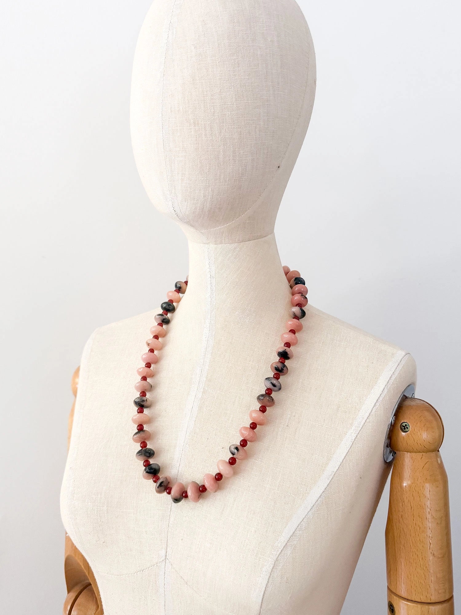 A beaded necklace featuring alternating light pink and grey marbled stone beads, dark red round beads, finished with an antique brass clasp.
