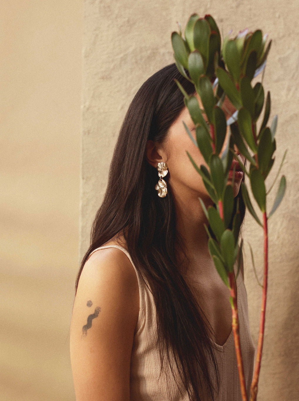 Woman with long dark hair holding a green plant wearing gold brass dangle earrings by Michelle Ross